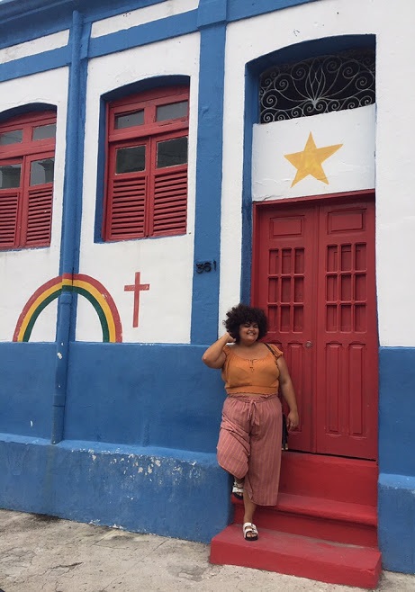 uiza em pé de cabelos cacheados curtos, blusa amarela e calça vermelha listrada. Atrás dela, uma casa em Olinda pintada com os símbolos da bandeira de Pernambuco.